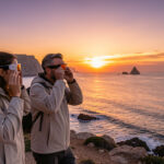 Deux femmes et un homme observent une éclipse solaire avec des lunettes de protection sur une côte espagnole au coucher du soleil.