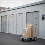 Où louer un box sécurisé, accessible tous les jours à Paris ? Vue réaliste d’un centre de self-stockage moderne près de Paris, avec une façade sécurisée, un accès contrôlé, quelques cartons et un chariot à l’entrée, éclairé par la lumière naturelle.