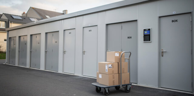 Vue réaliste d’un centre de self-stockage moderne près de Paris, avec une façade sécurisée, un accès contrôlé, quelques cartons et un chariot à l’entrée, éclairé par la lumière naturelle.