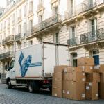 Camion de déménagement garé dans une rue parisienne, entouré de cartons empilés devant des immeubles haussmanniens baignés d’une lumière douce.
