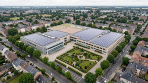 Vue aérienne réaliste d’un groupe scolaire et d’un gymnase à Arras, avec des toitures couvertes de panneaux solaires dans un environnement urbain du Nord de la France, sous une lumière douce.